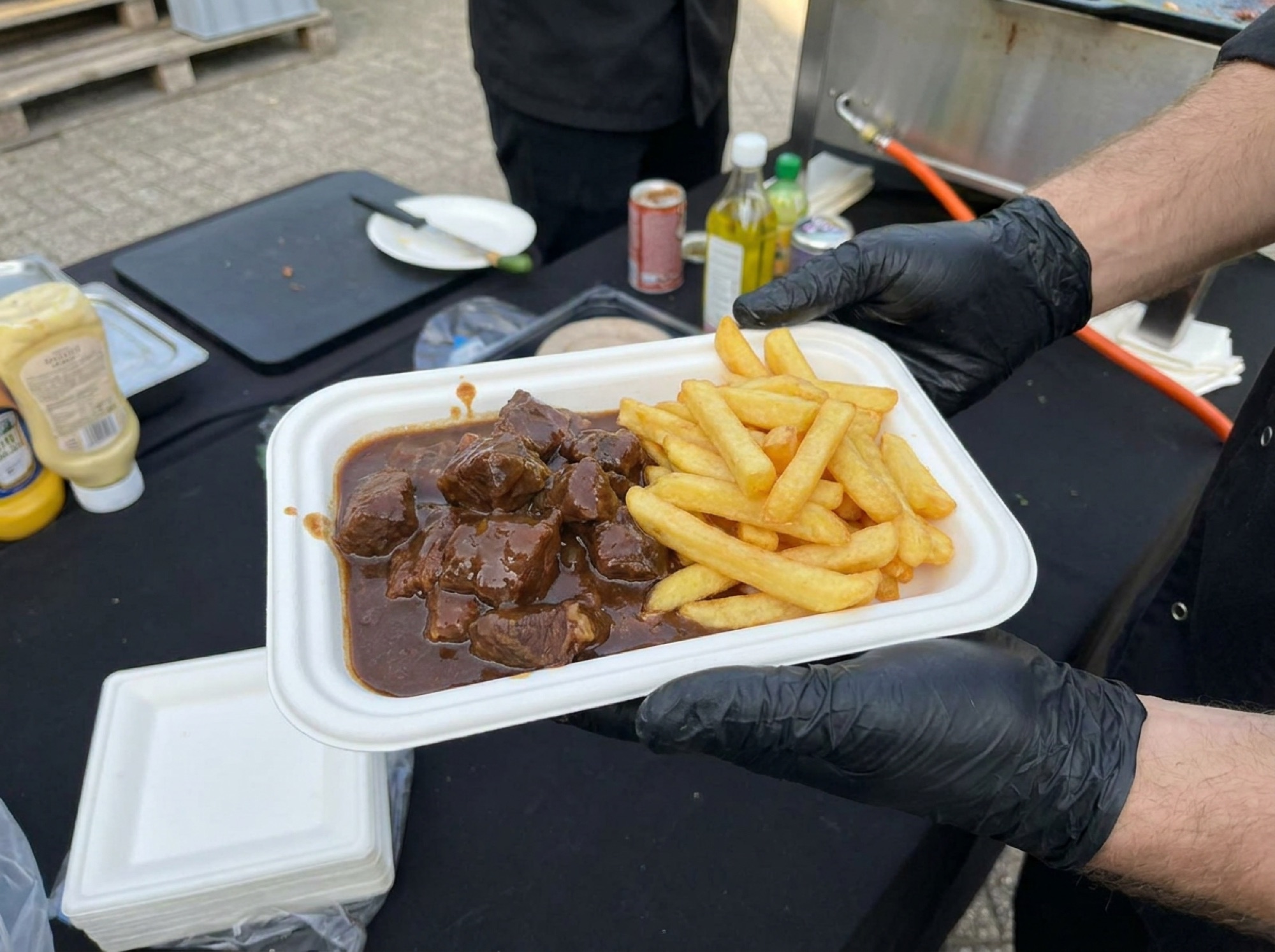 Stoofvlees met frieten in wit suikerrietbakje, vastgehouden met zwarte handschoenen aan de cateringcounter